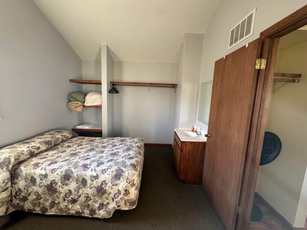 A small, compact room with a bed, a wooden wardrobe, a small table, and a mirror.