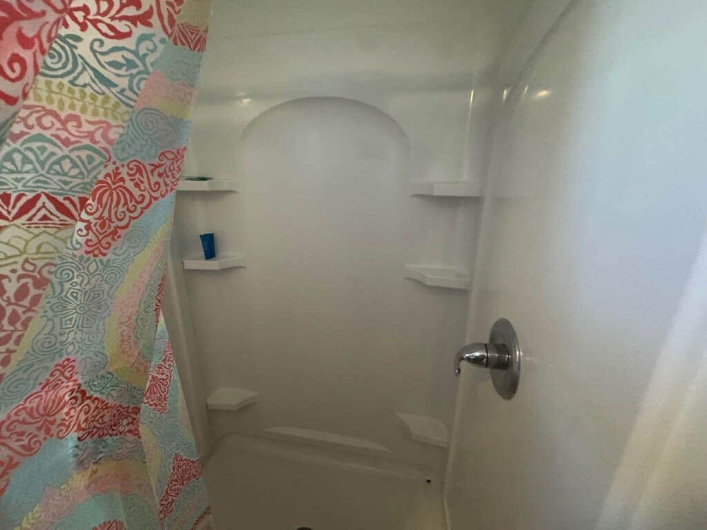 Een badkamer met een douche, toilet en wastafel. Boven de douche bevinden zich planken en een deur met een handgreep.
