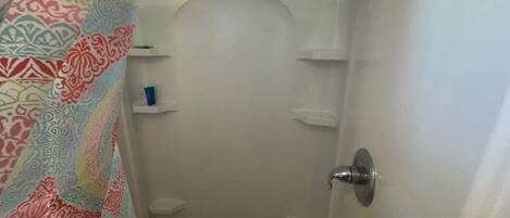 Een badkamer met een douche, toilet en wastafel. Boven de douche bevinden zich planken en een deur met een handgreep.