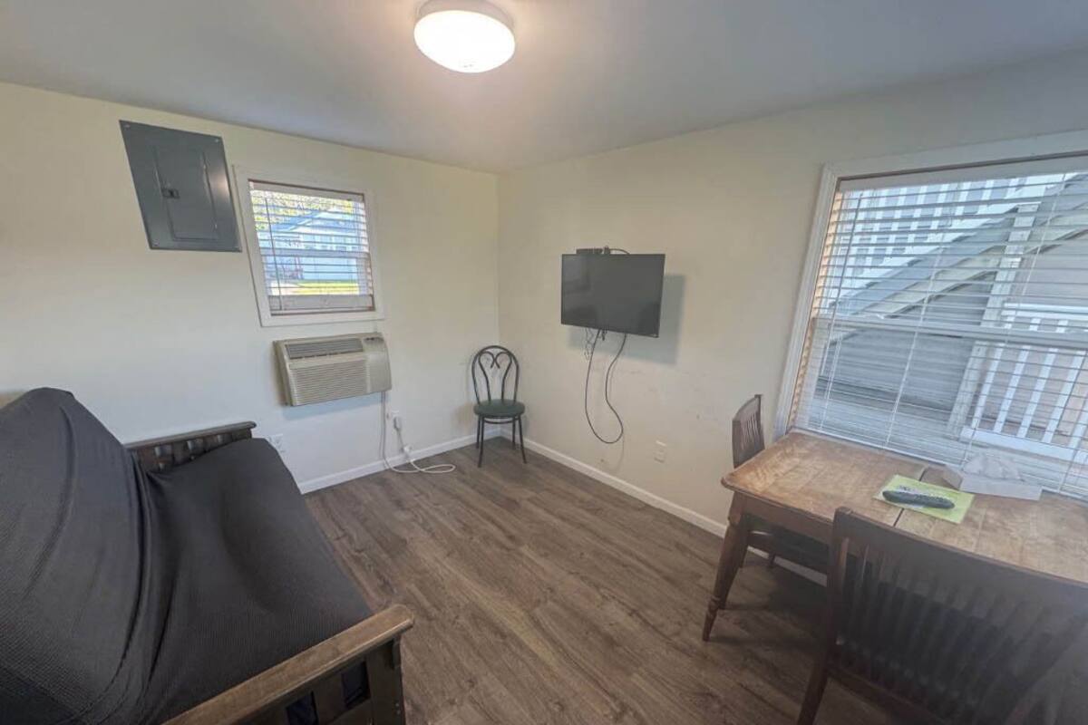 Une pièce comprenant un lit, une table à manger avec des chaises, un téléviseur fixé au mur et deux fenêtres équipées de stores.