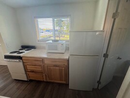 Cocina compacta con estufa, microondas y refrigerador, armarios de madera y una ventana con persianas.
