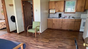 Una stanza compatta con pavimento in legno, un letto, una sedia verde, un angolo cottura con mobili in legno e un forno a microonde.