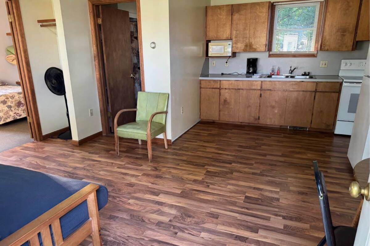 Une pièce de taille modeste avec un plancher en bois, un lit, une chaise verte et une petite cuisine équipée de meubles en bois et d’un four à micro-ondes.
