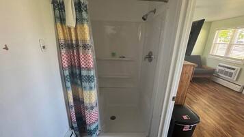 A walk-in shower with a patterned shower curtain, a wooden floor, and a window with blinds.