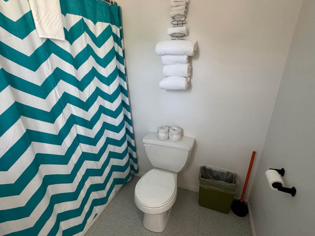 A bathroom with a white toilet, a green and white chevron shower curtain, a towel rack with folded towels, a trash bin, and a broom.