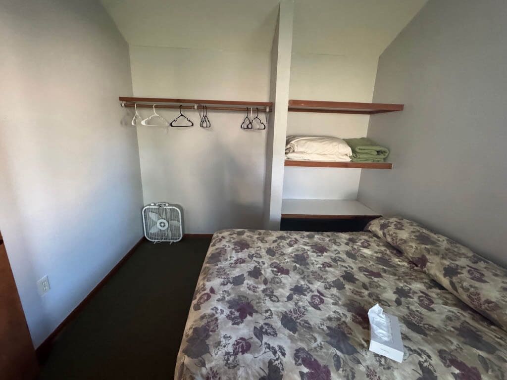 Habitación pequeña con una cama, un ventilador y un estante con ropa doblada.