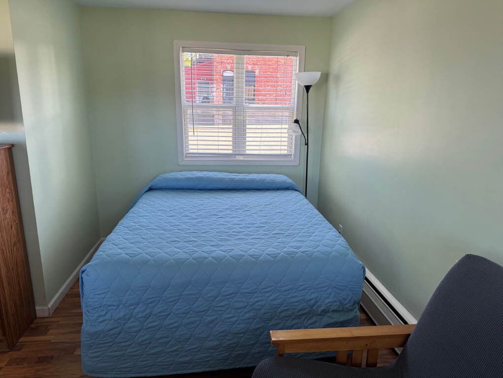 A single bed with a blue quilt, a nightstand, a lamp, and a chair in a room with light green walls and a window with blinds.