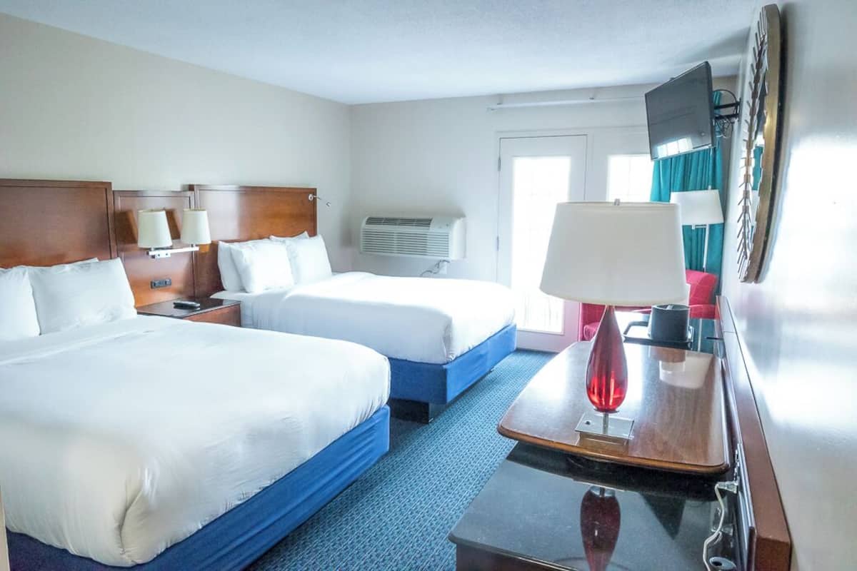 A hotel room with two beds, a desk, a TV, and a wall-mounted air conditioning unit.