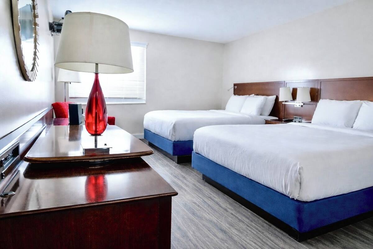 A hotel room with two beds, a desk, a lamp, and a window.