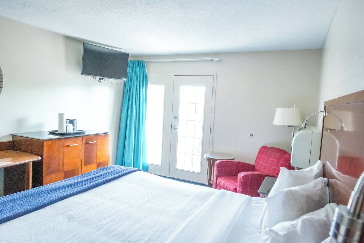A hotel room with a bed, a desk, a chair, a TV mounted on the wall, and a door with a window.