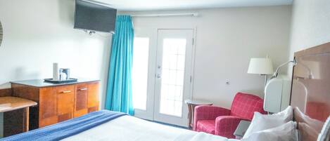 A hotel room with a bed, a desk, a chair, a TV mounted on the wall, and a door with a window.