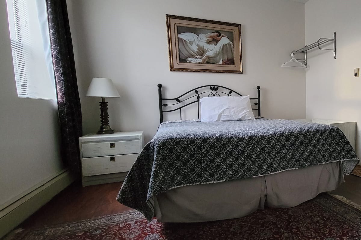 A bedroom with a bed, a nightstand, a lamp, a painting, and a window with curtains.