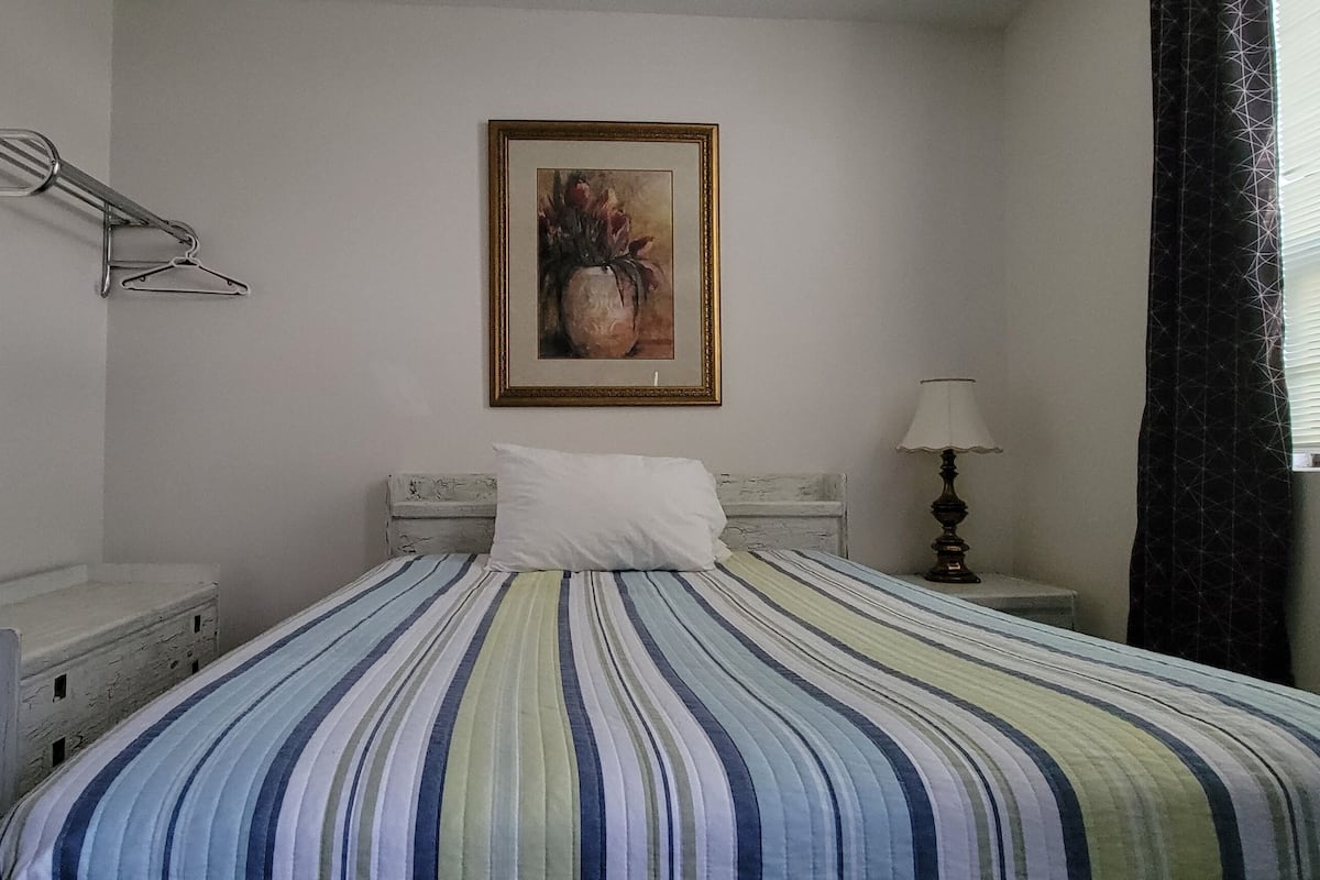 A bed with a striped bedspread, a bedside lamp, a framed picture on the wall, and a window with curtains.