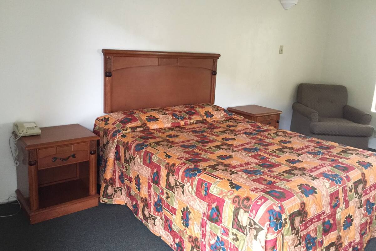 A single bed with a floral bedspread, a wooden nightstand with a telephone, and a chair against the wall in a simple room.