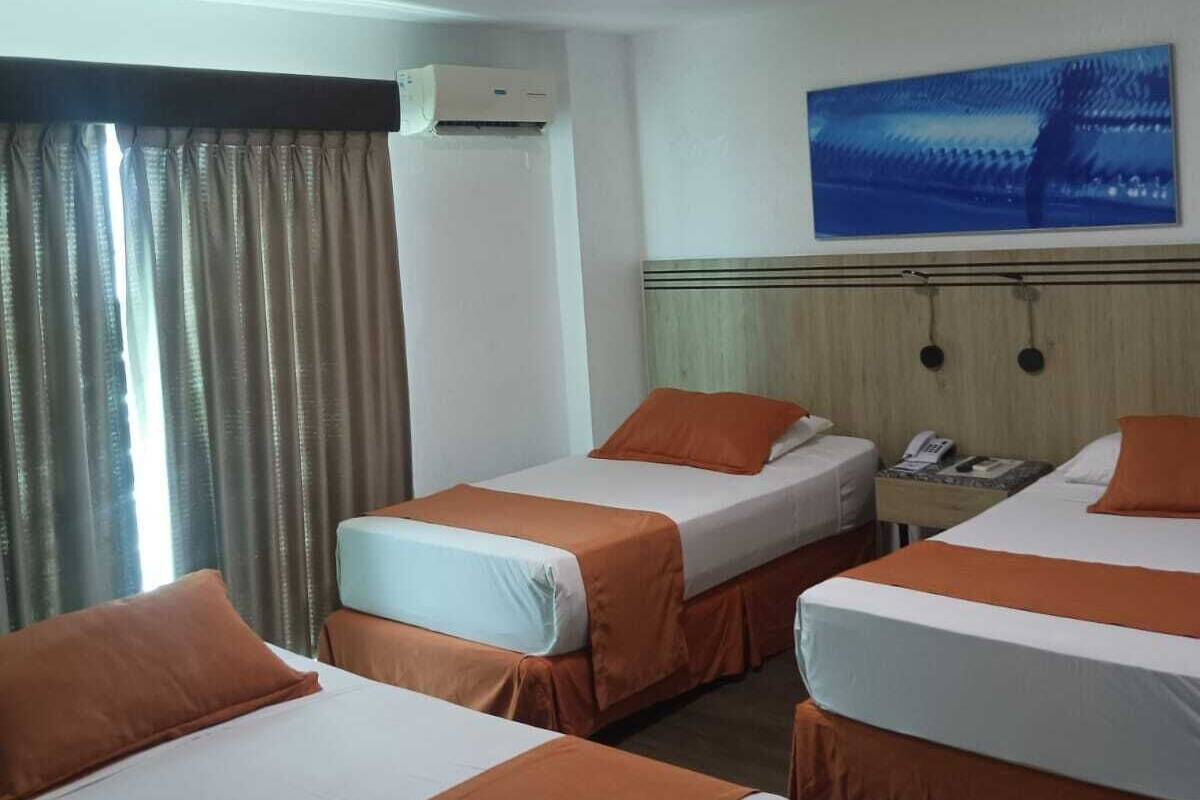 A hotel room with three beds, each with orange bedding, a large window with curtains, an air conditioning unit, a wall-mounted phone, and a painting above the beds.