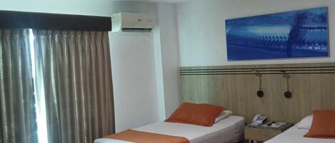 Habitación de hotel con tres camas, cada una con ropa de cama naranja, un ventanal con cortinas, un equipo de aire acondicionado, un teléfono en la pared y un cuadro sobre las camas.
