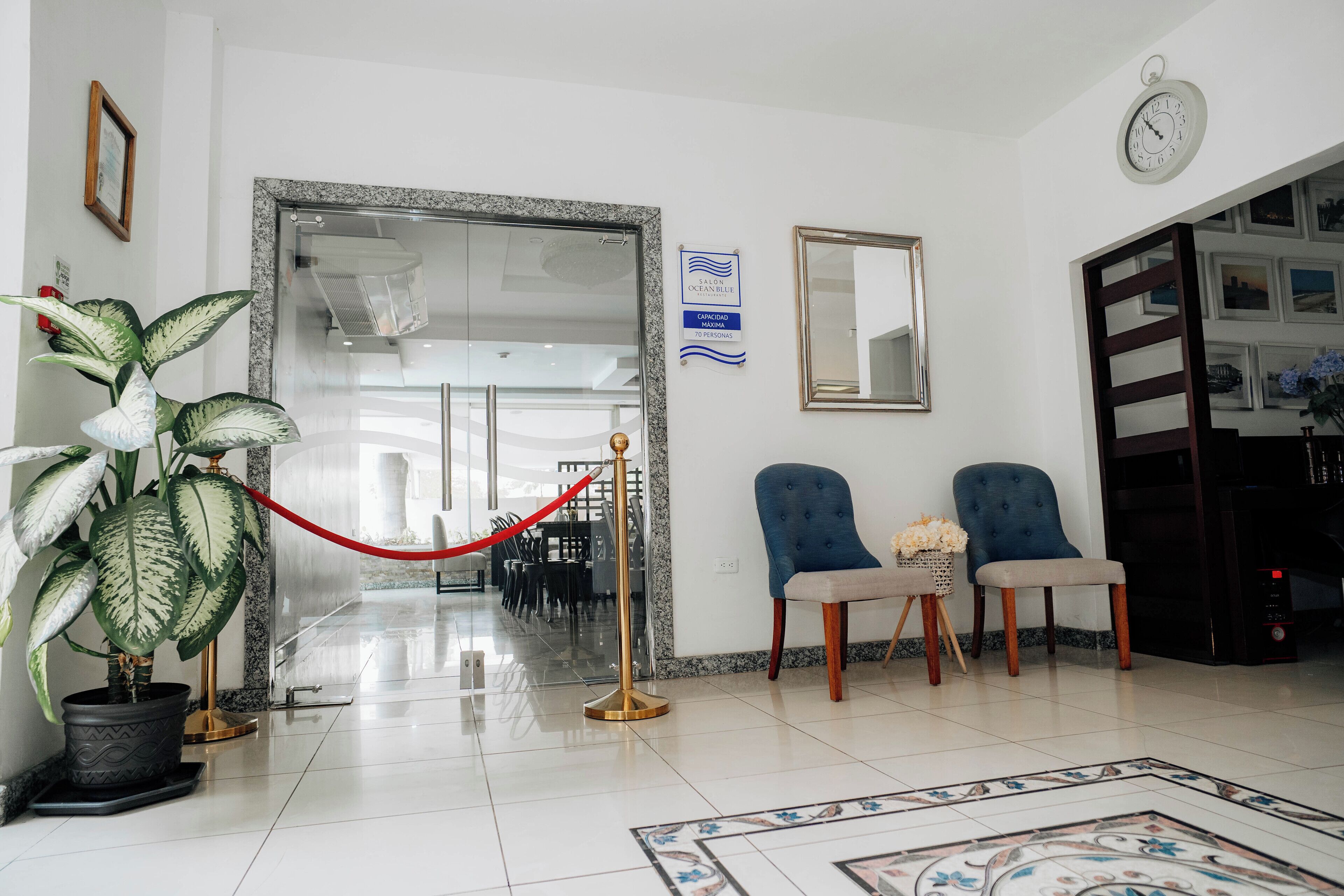 Un vestíbulo de hotel con mostrador de recepción, dos sillones, una planta en maceta, una barrera de cuerda roja y un reloj en la pared.