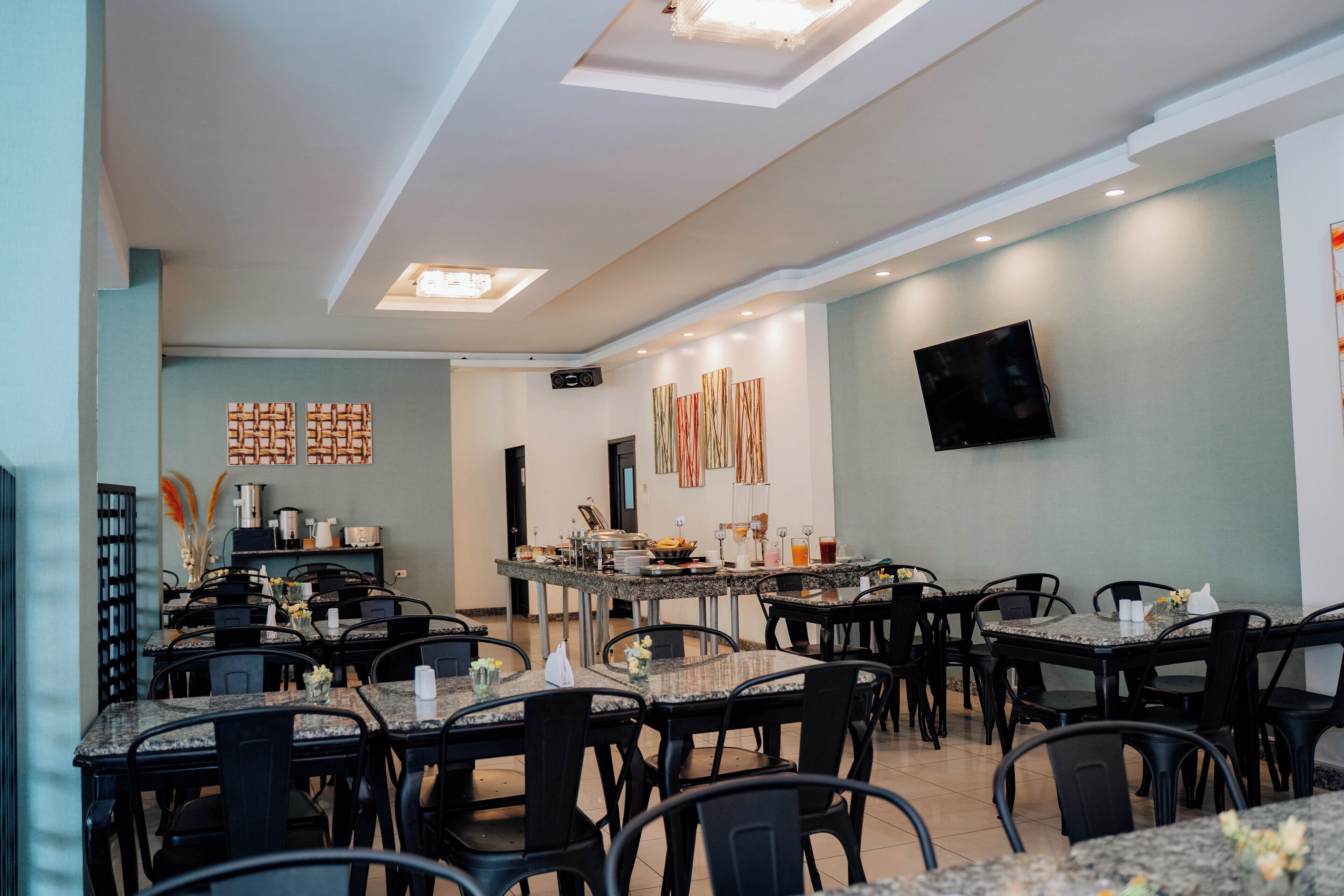 A restaurant with multiple tables and chairs, a kitchen area, and a flat-screen TV mounted on the wall.