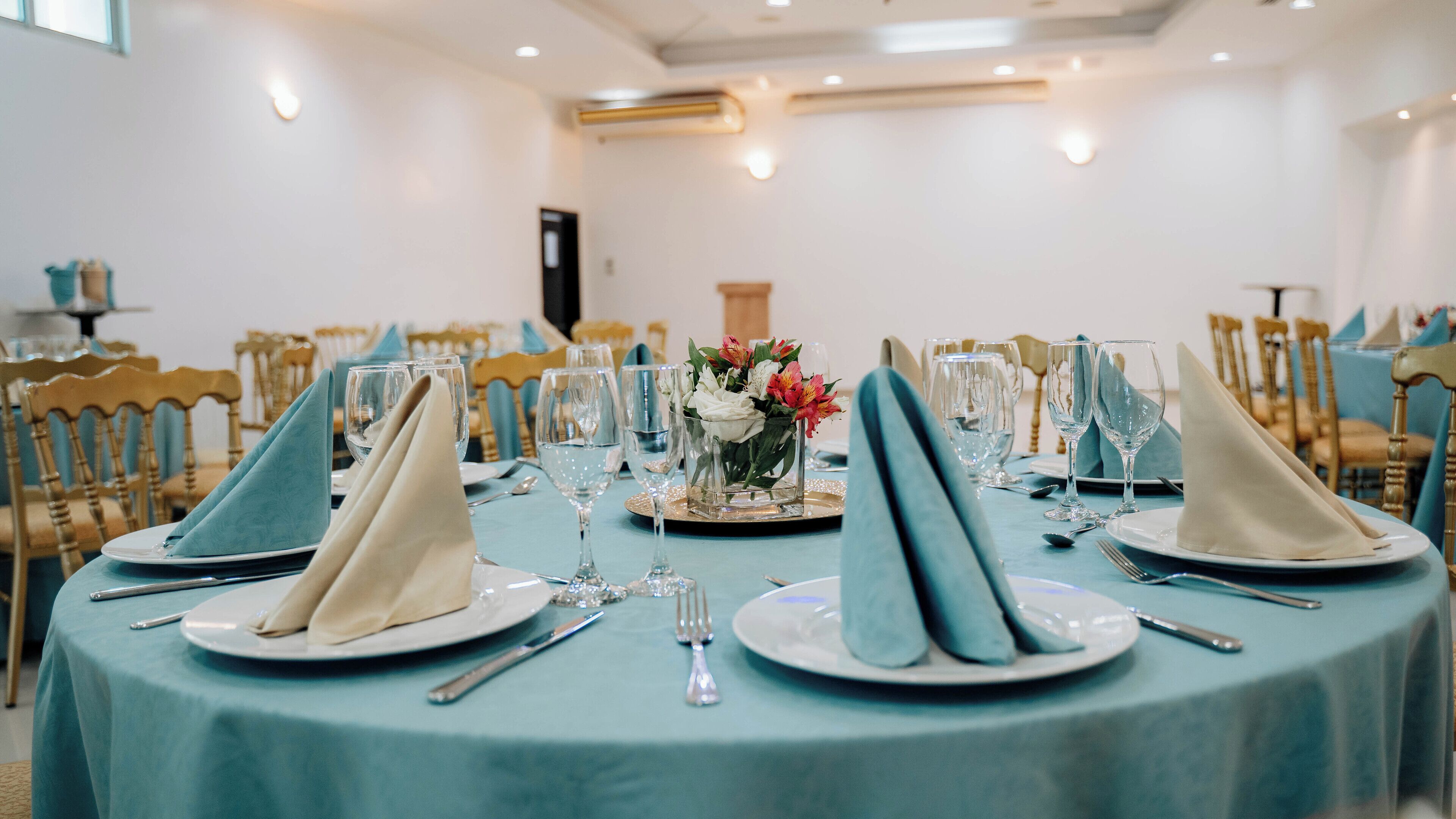 A formal dining room with round tables set for a meal, featuring blue tablecloths, white plates, and clear glassware.