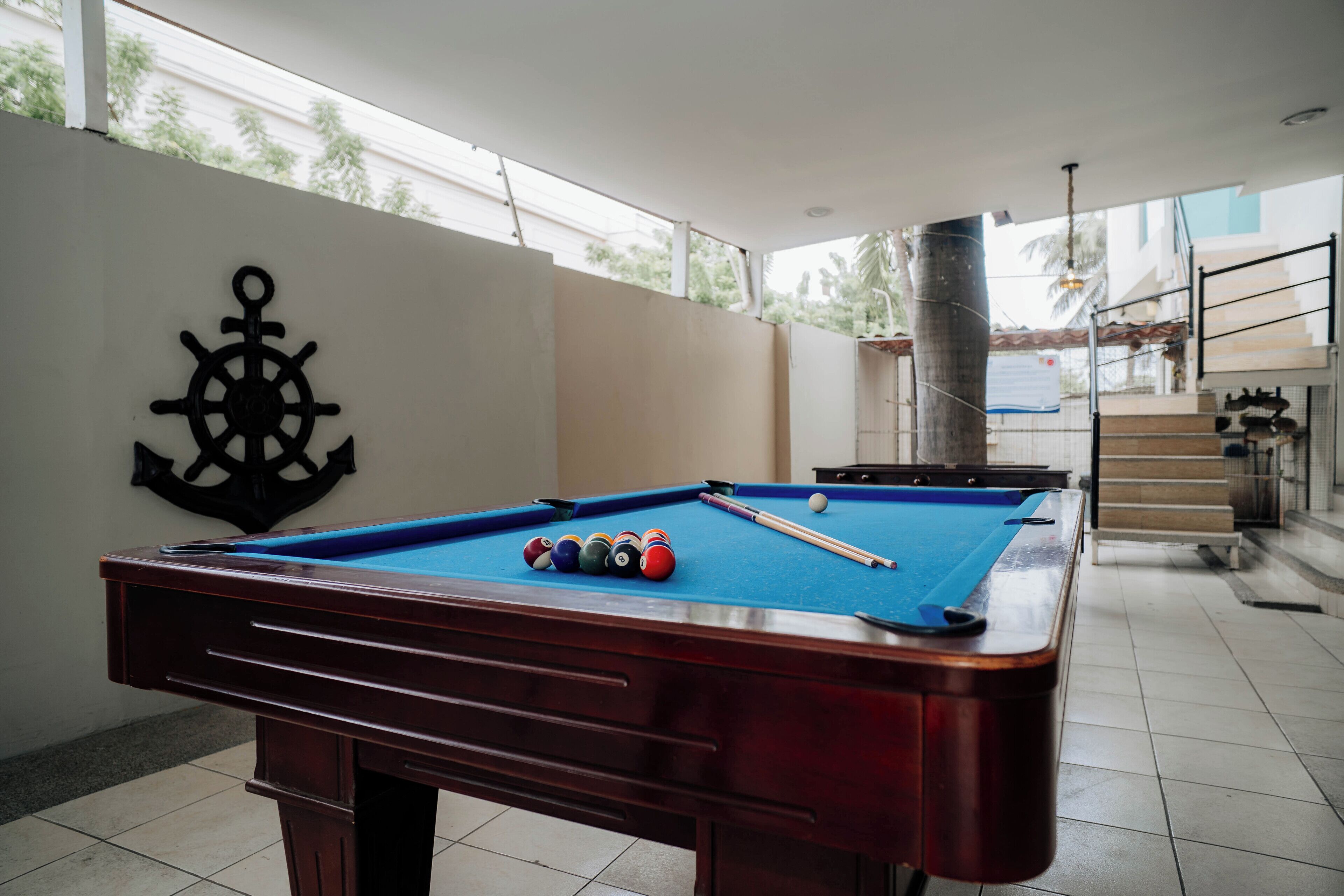 A pool table with balls and a cue stick, a wall with a ship's wheel decoration, and a staircase in the background.