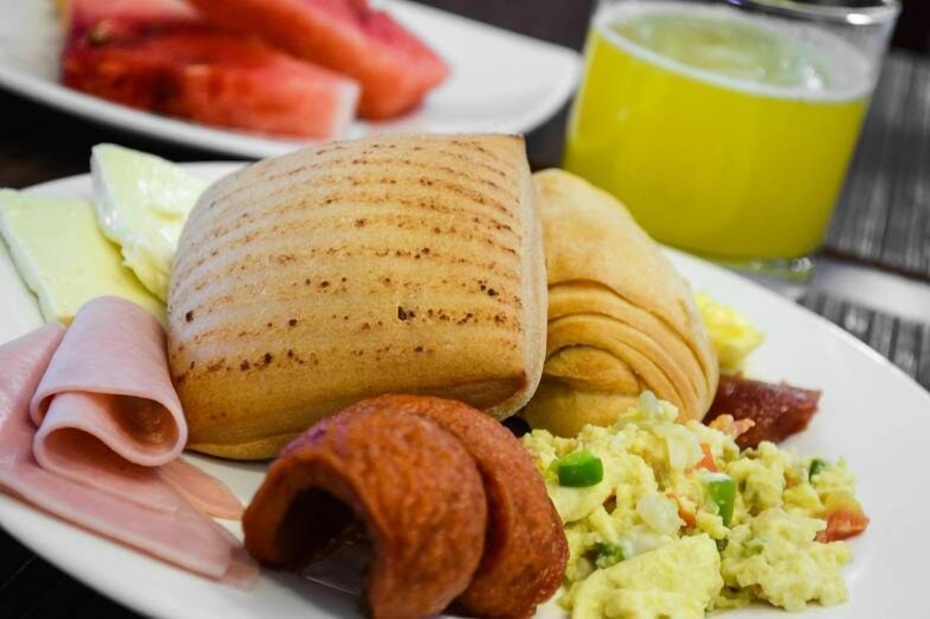 A breakfast plate with a rolled pastry, ham, a croissant, scrambled eggs, and a glass of yellow juice.