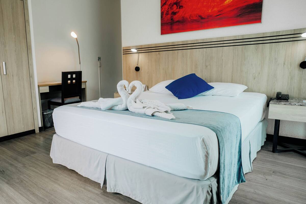 A hotel room with a bed, a desk, a chair, a wardrobe, and a painting on the wall.