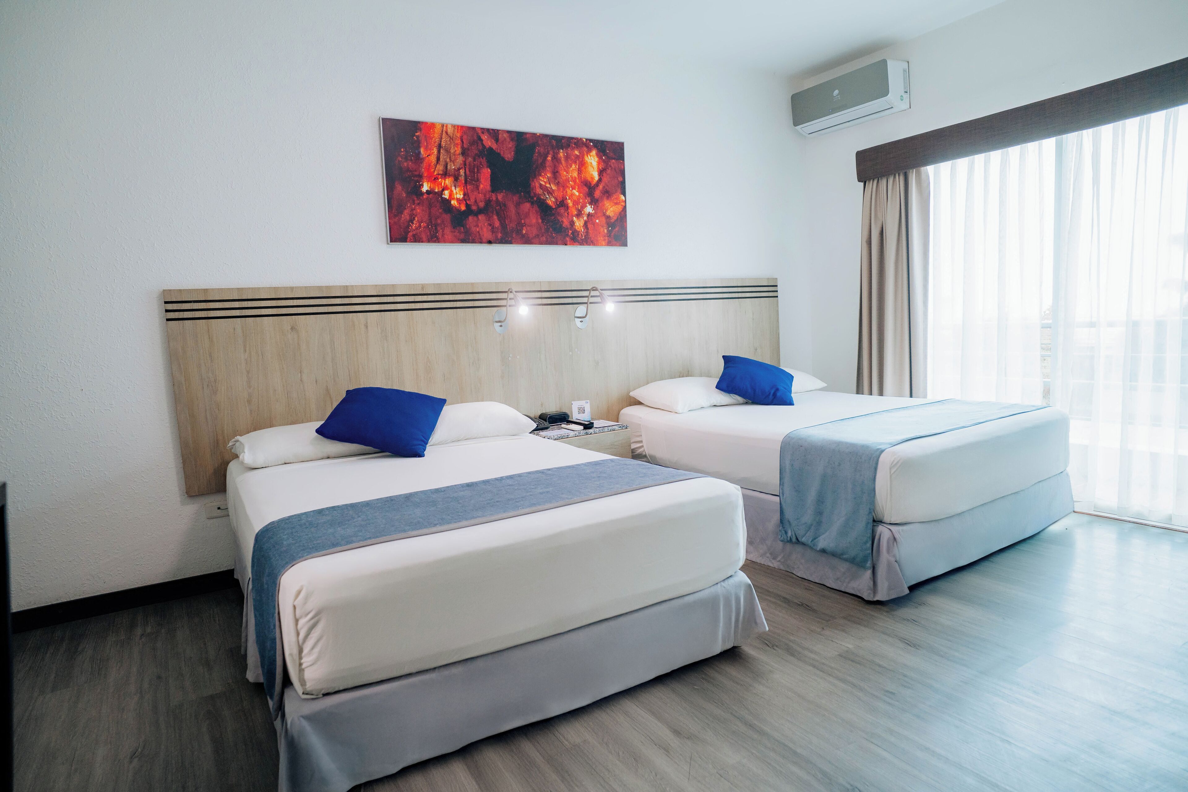 A hotel room with two beds, a wooden headboard, a painting, and a window with curtains.