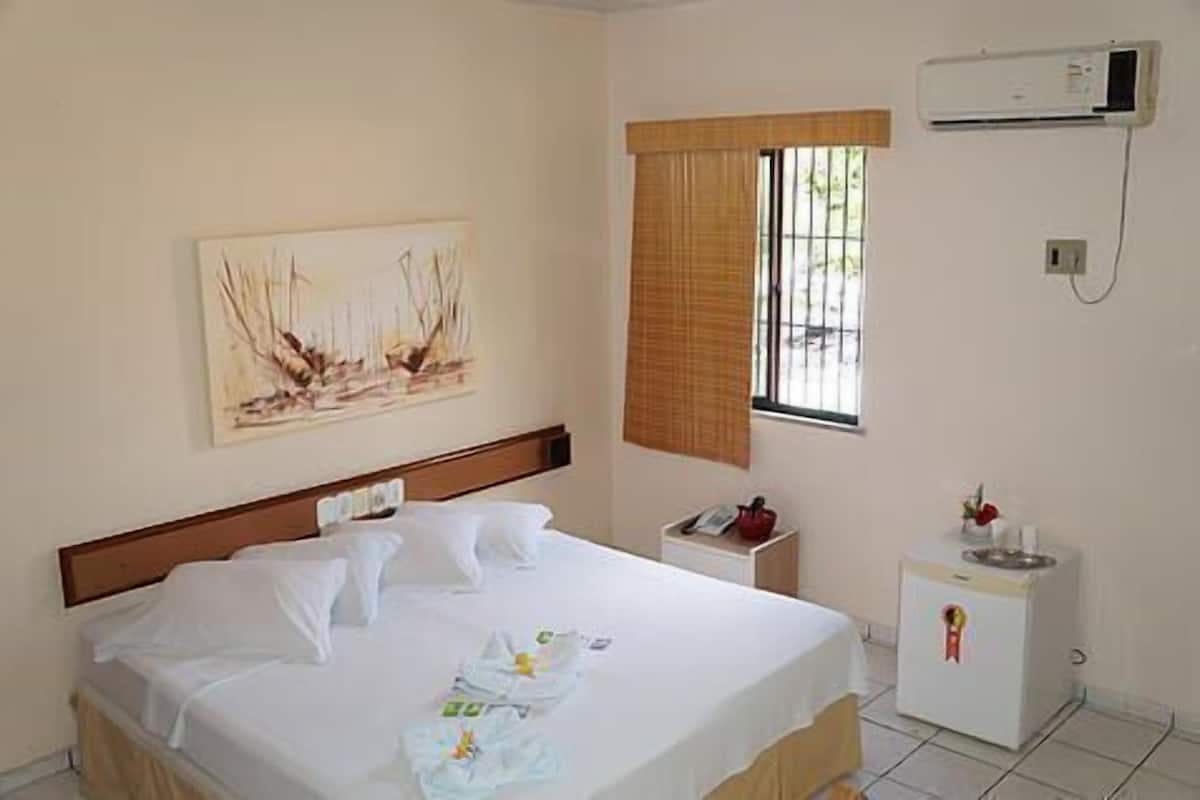 Une chambre d’hôtel comprenant un lit, une petite table de chevet, un réfrigérateur, une fenêtre avec des stores et un appareil de climatisation.