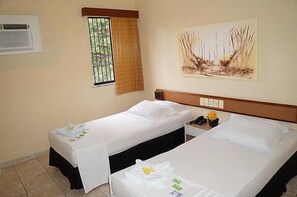 A hotel room with two beds, a window with bars, a painting on the wall, and a nightstand with a phone and a vase of flowers. - Hotel Malibu (Fortaleza)