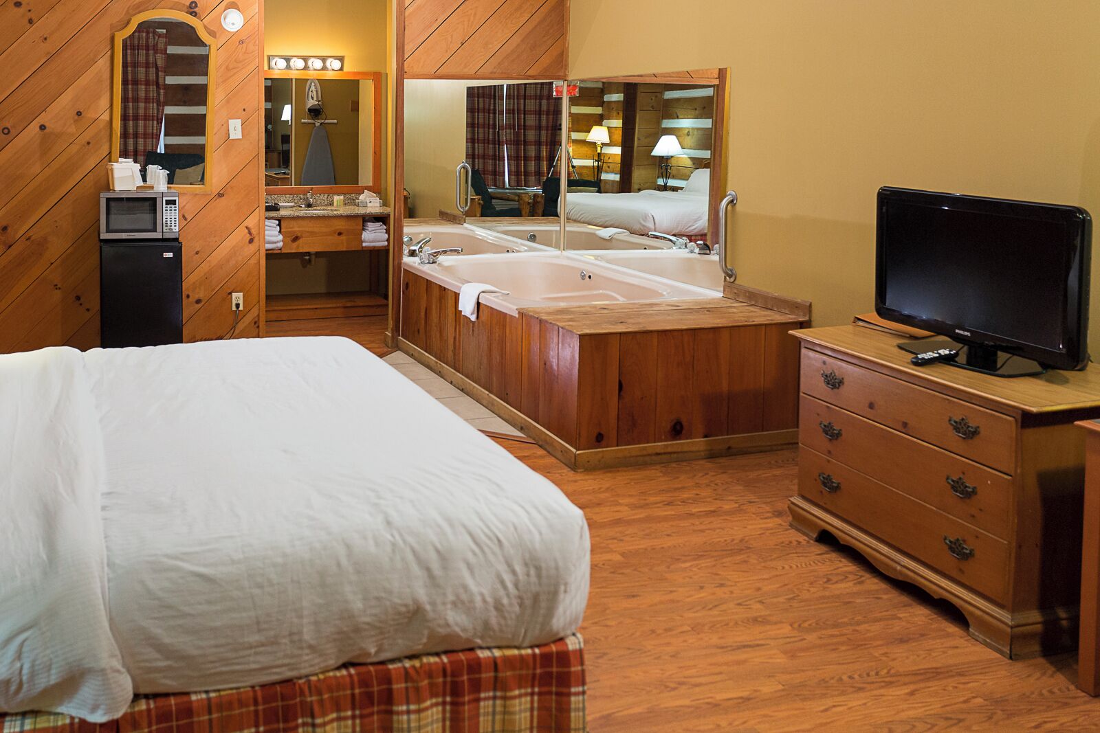 Una habitación de hotel con cama, cómoda de madera, televisor, baño con bañera y espejo.