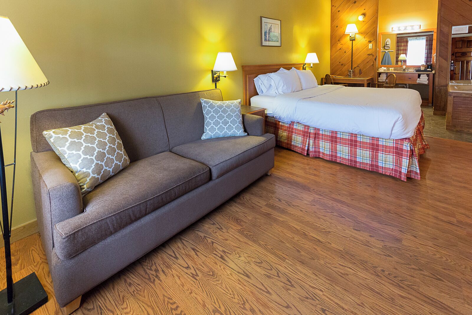 A room with a sofa, a bed with a plaid bedspread, a desk with a chair, and wall-mounted lamps.