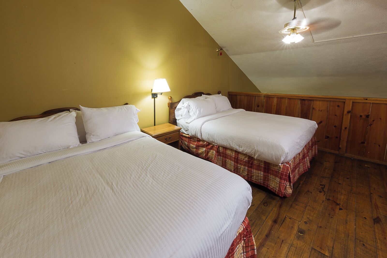 A room with two beds, wooden walls, a ceiling fan, and a nightstand with a lamp.