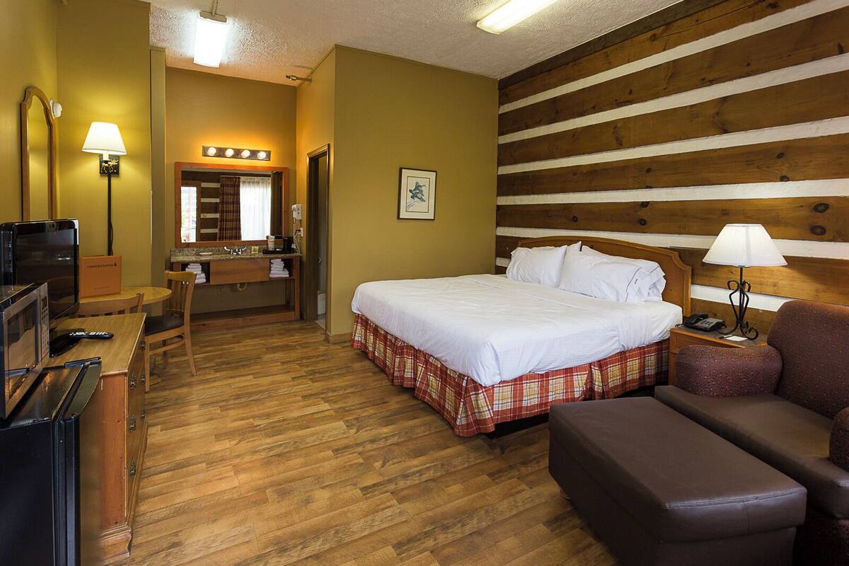 A hotel room with a bed, a desk with a mirror, a chair, a couch, and a wooden wall.