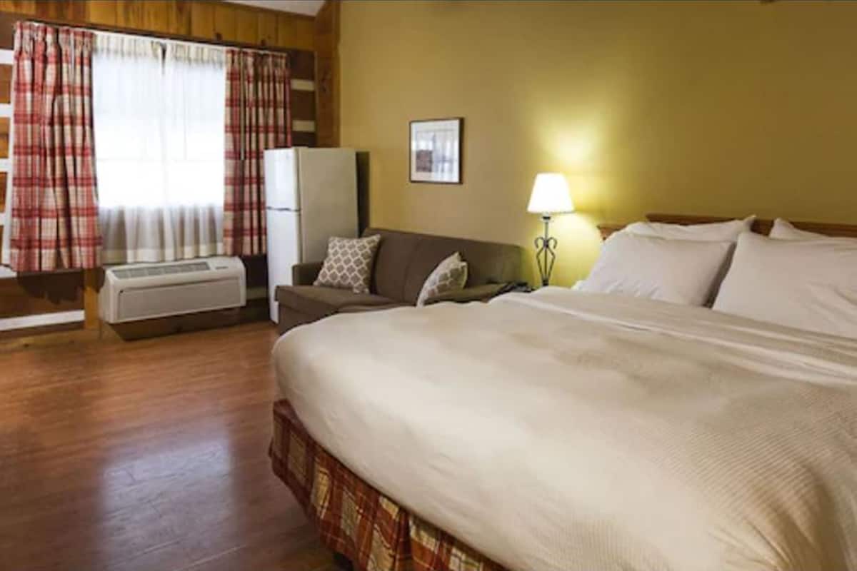 A hotel room with a large bed, a sofa, a small table, a lamp, a refrigerator, and an air conditioning unit.