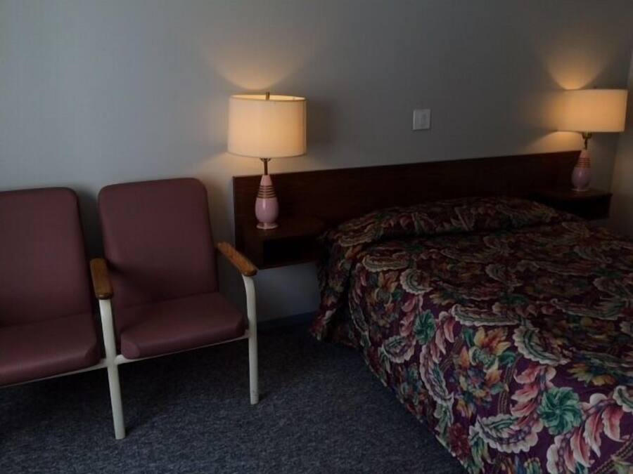 Une chambre d’hôtel avec deux chaises, un lit avec une literie à motifs floraux et deux lampes de chevet.