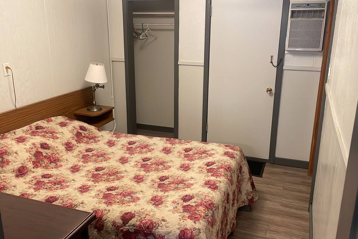 A hotel room with a bed, a nightstand, a lamp, a wardrobe, and an air conditioning unit.