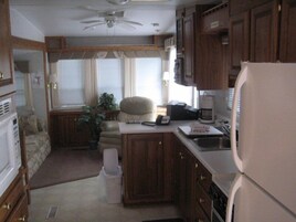 Cocina compacta para camper con armarios de madera, un refrigerador, una cafetera y un comedor con una silla y mesa.