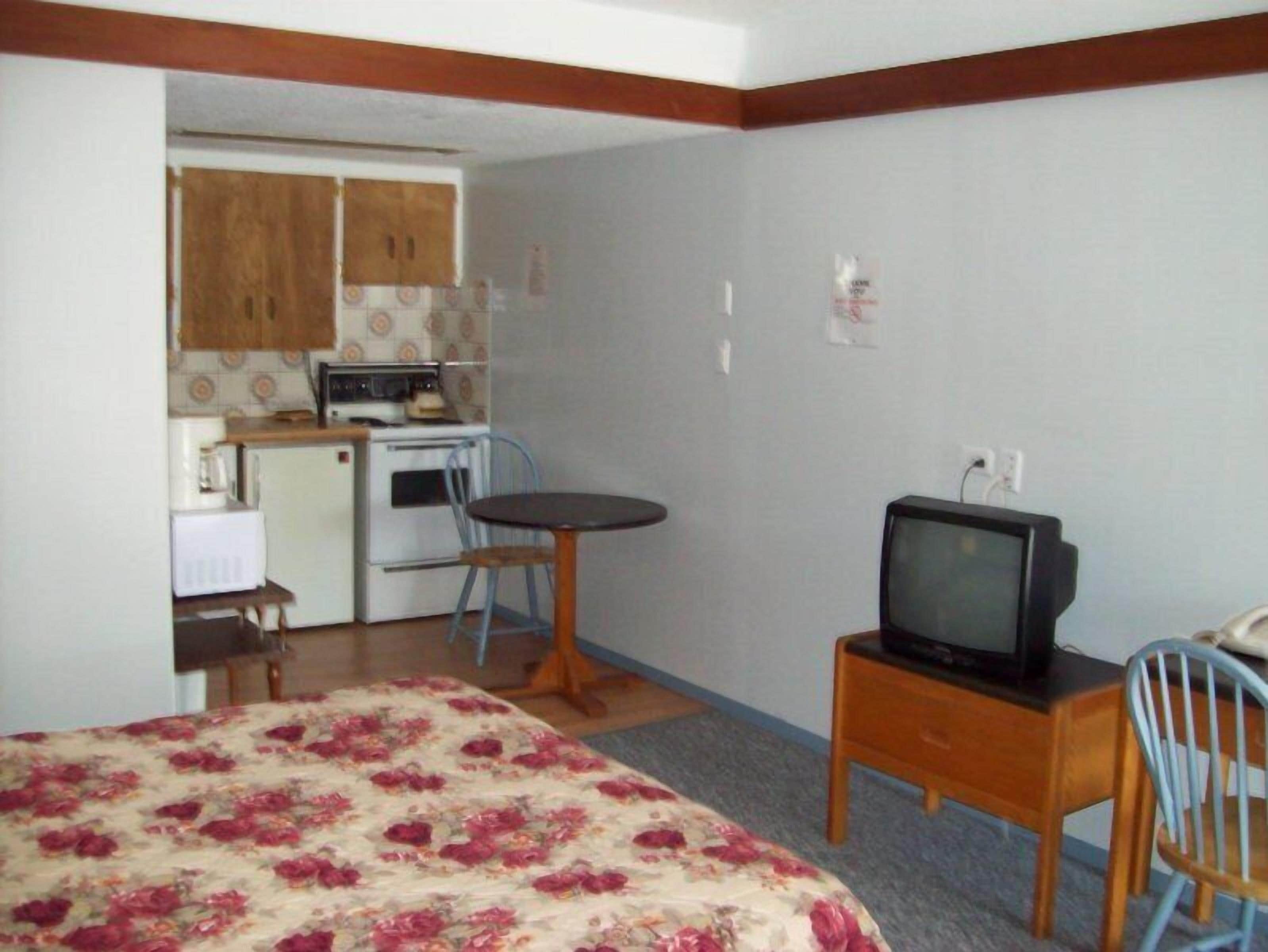Una habitación compacta con cama, una mesita con televisor, una kitchenette equipada con estufa y microondas, y una silla.