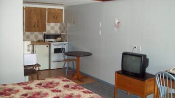 Una habitación compacta con cama, una mesita con televisor, una kitchenette equipada con estufa y microondas, y una silla.