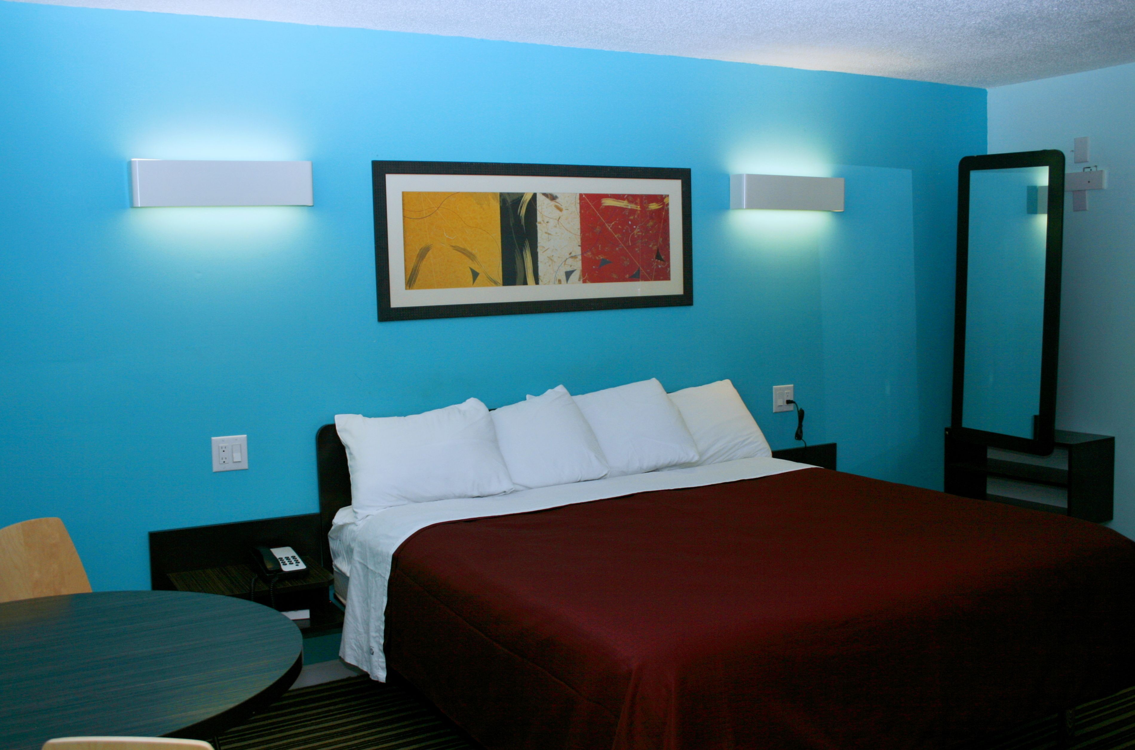 Habitación de hotel con cama, mesita de noche, teléfono, cuadro abstracto enmarcado y espejo.