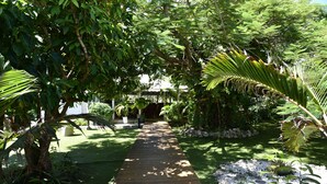 A wooden pathway leading through a lush garden with various plants and a small bridge in the distance. - Hotel Le Soleil Levant (Capesterre-de-Marie-Galante)