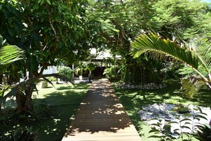 A wooden pathway leading through a lush garden with various plants and a small bridge in the distance.