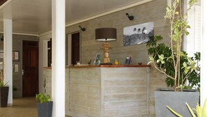 A reception area with a wooden reception desk, a lamp, a framed picture on the wall, and potted plants. - Hotel Le Soleil Levant (Capesterre-de-Marie-Galante)