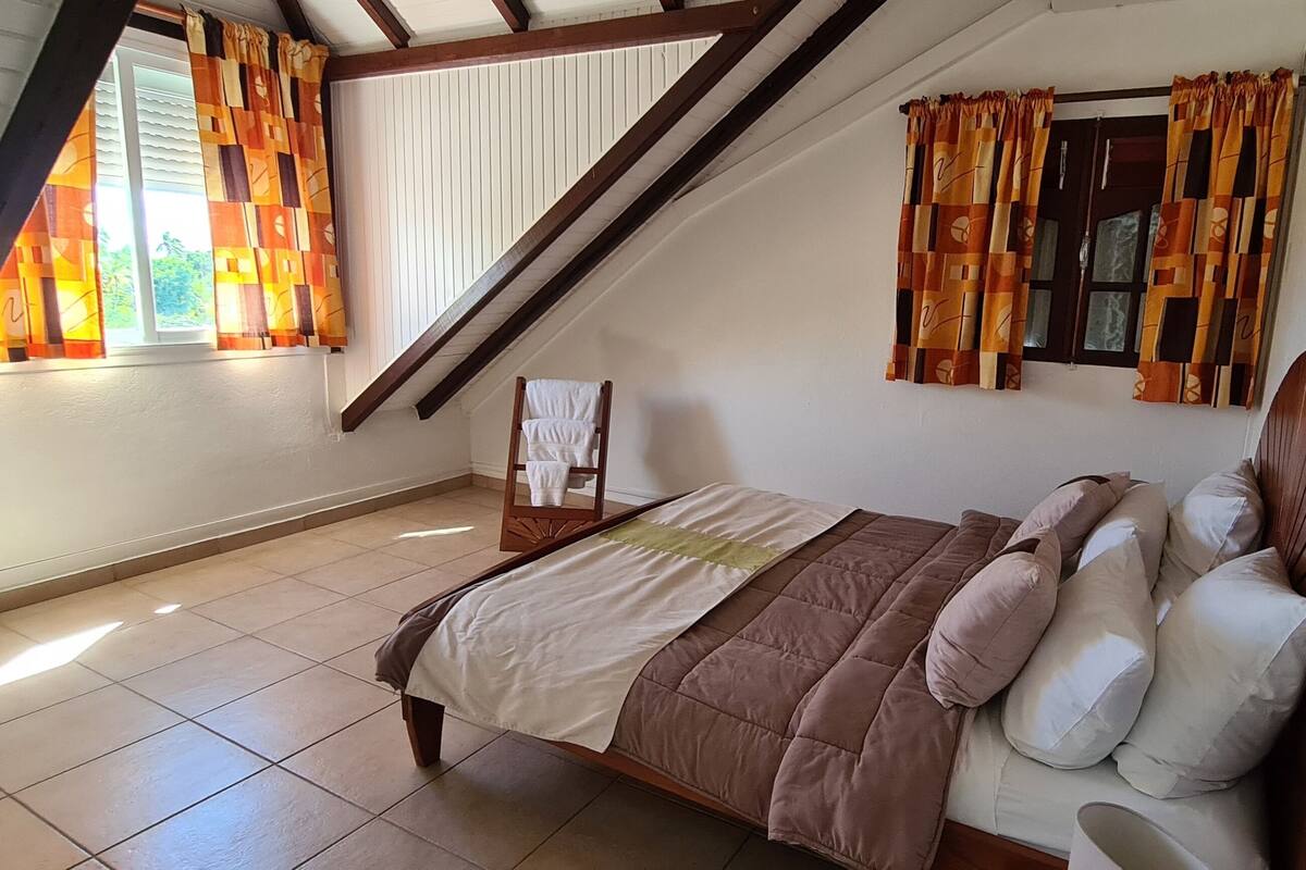 A bedroom with a sloped ceiling, a bed with a brown quilt, white pillows, and a wooden headboard. There is a window with curtains, a small table, and a door with a handle.