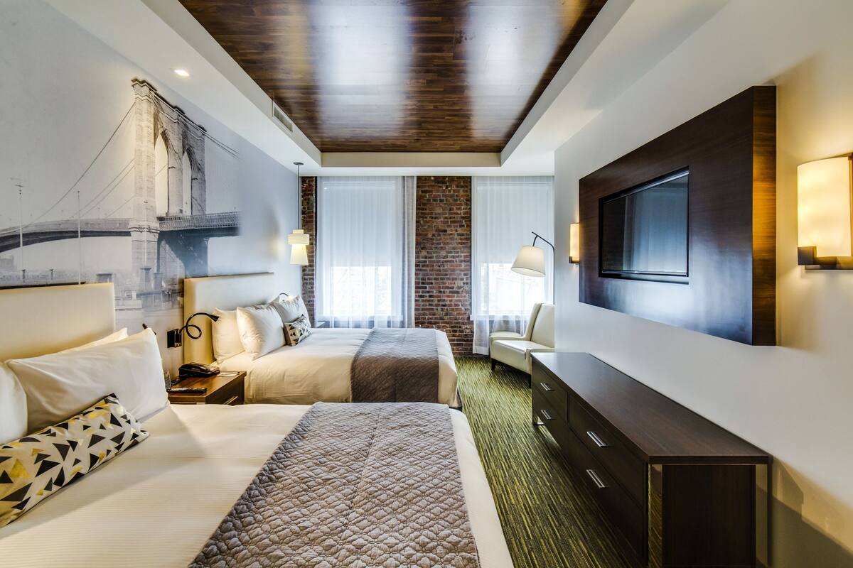 A hotel room with two beds, a large TV, a brick accent wall, and a view of a bridge.