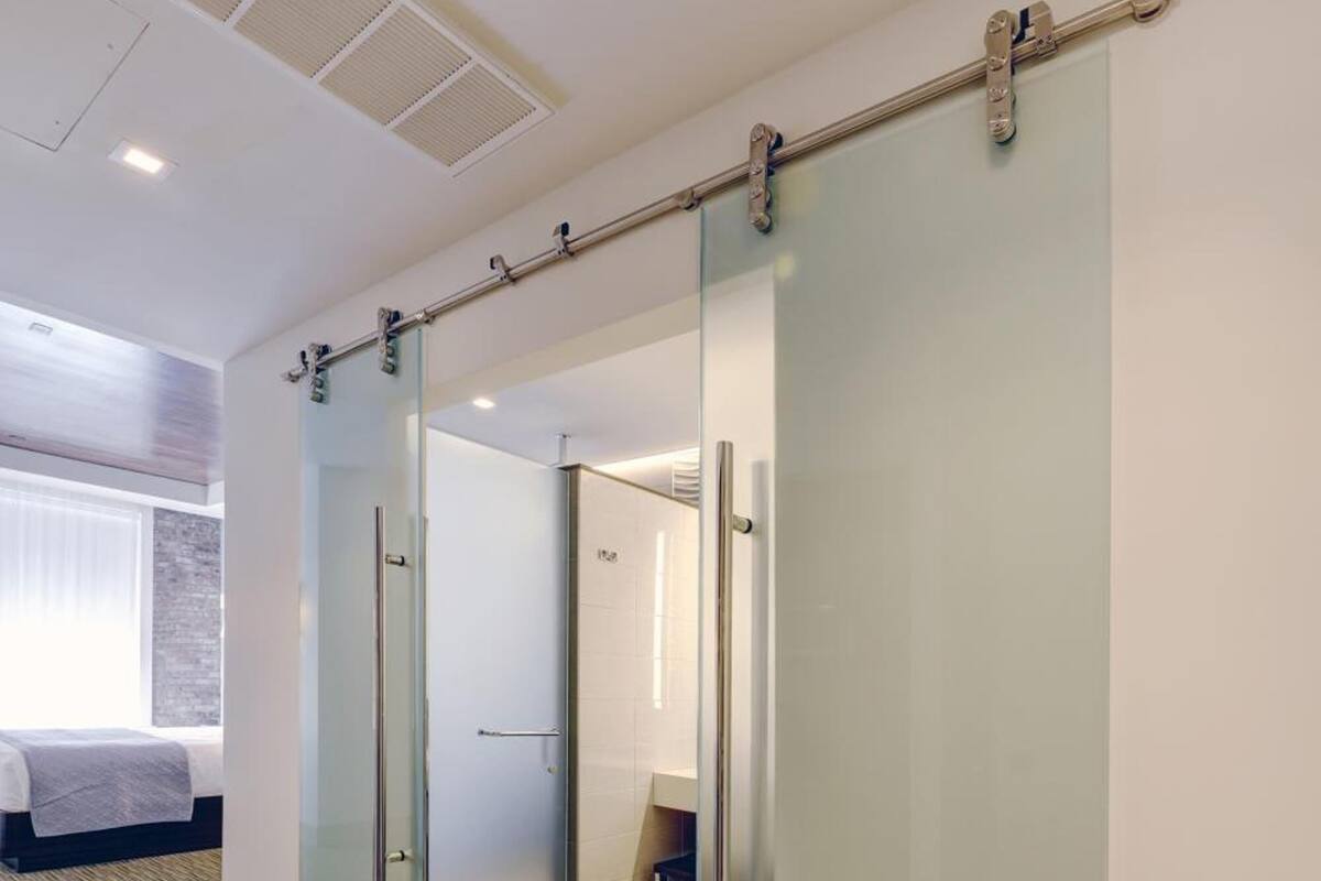 A hotel room with a sliding door leading to a bathroom, a bed with a white headboard, and a window with blinds.