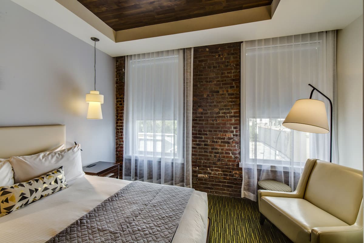 A hotel room with a bed, a chair, a lamp, and a view through sheer curtains.