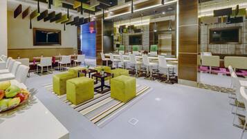 A modern hotel lobby with a central seating area, a reception desk, and a large TV screen.