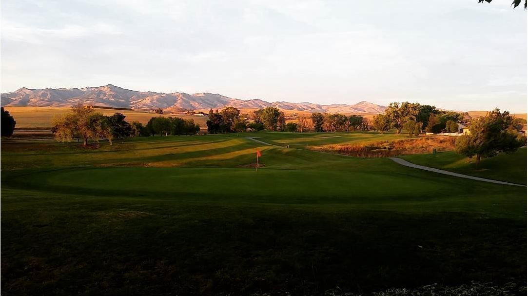 Um campo de golfe com uma bandeira, árvores e montanhas ao fundo.