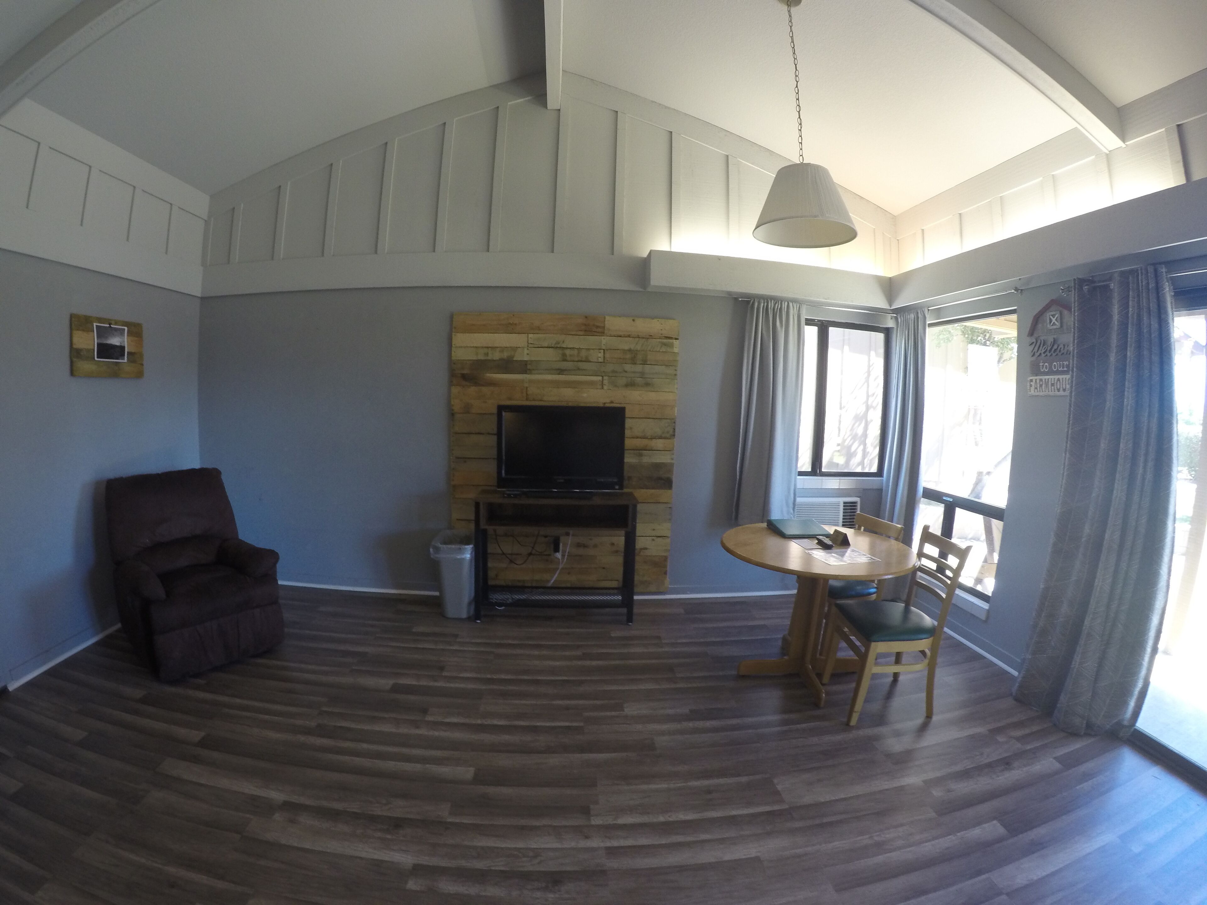 Una habitación amplia con suelo de madera, un televisor montado en una pared de madera, una mesa de comedor con sillas, un sillón marrón y un ventanal con cortinas.